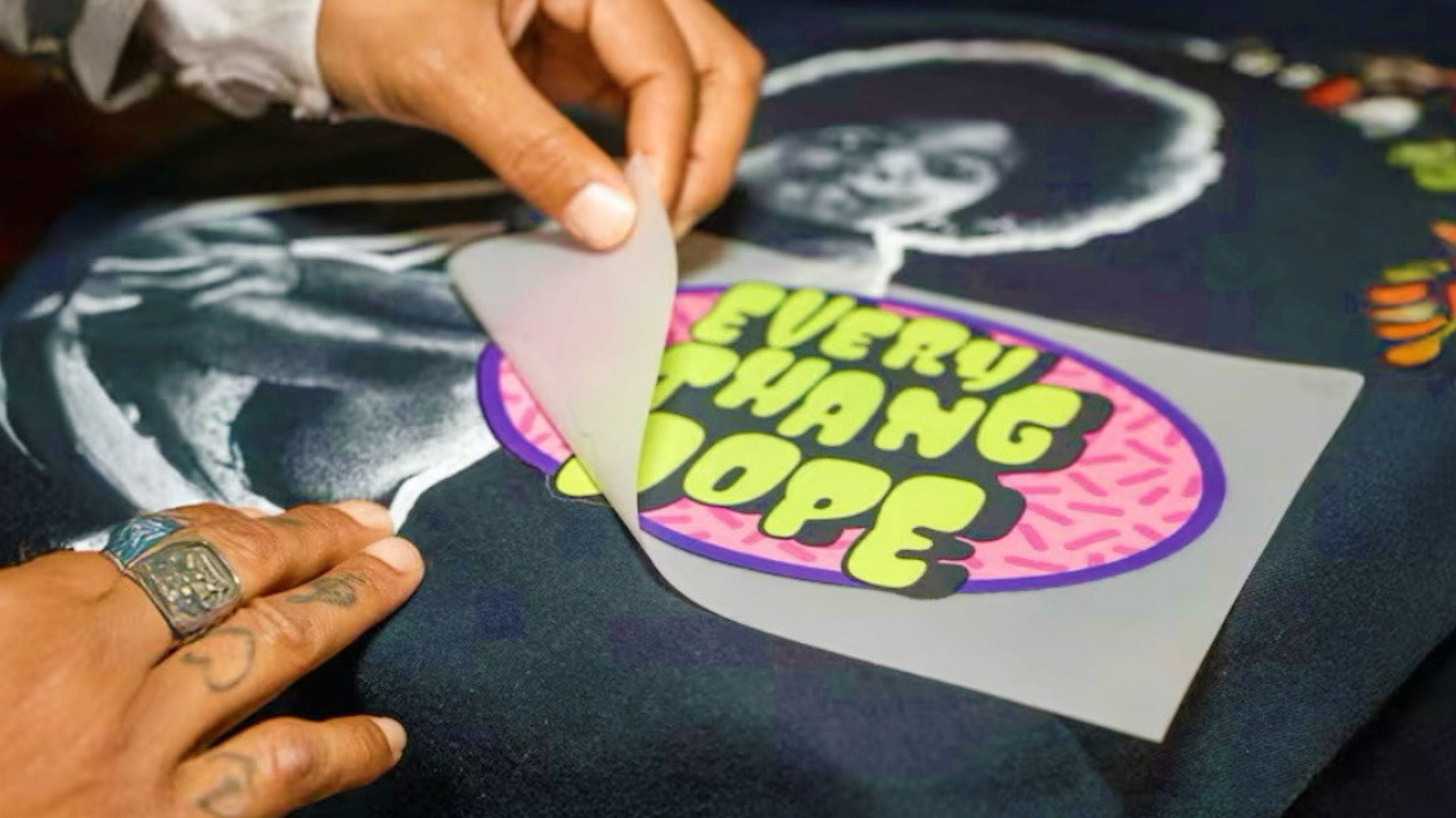 Hands applying a custom Everythang Dope retro vinyl transfer graphic onto a black t-shirt in a textile workshop.