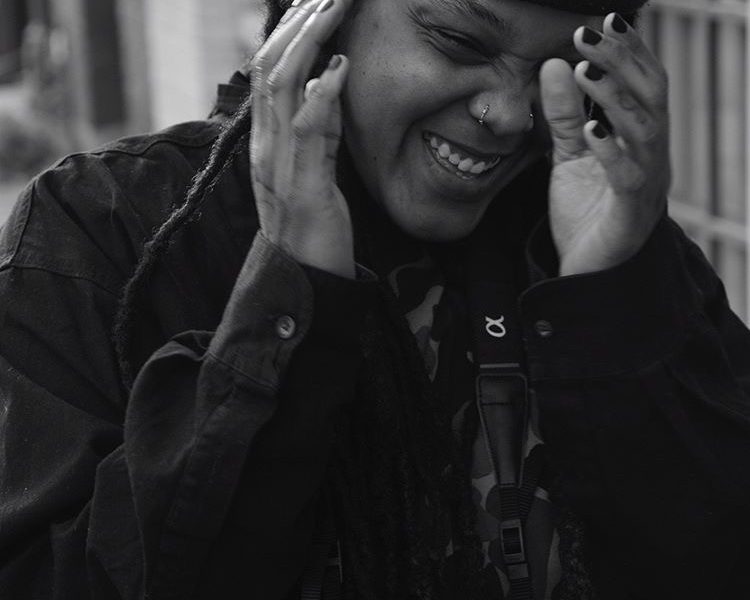 A black and white close-up photo of Aaliyah Arrington wearing a dark jacket and a beanie, holding their face in laughter with hands near their ears.