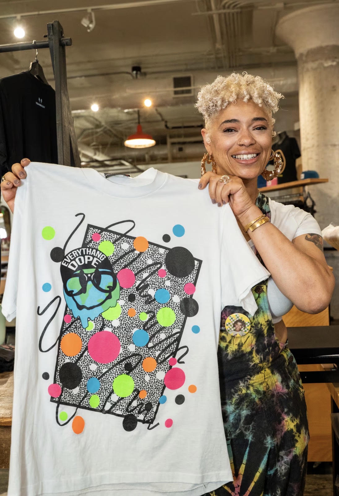 Everythang Dope founder holding up a custom 90s retro graphic t-shirt featuring a colorful graffiti design with the text 'EVERYTHANG DOPE' and a cartoon face wearing sunglasses inside the store workshop