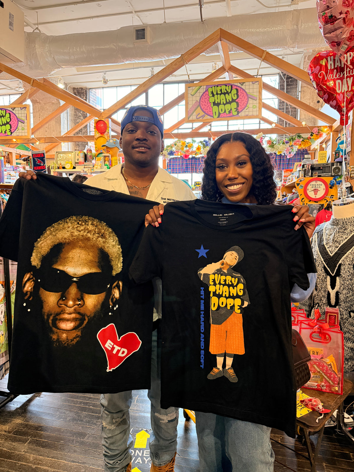 A man and a woman stand in a retail store holding up two black graphic T-shirts. The shirt on the left features a large portrait of a man wearing sunglasses with a red heart and the text 'ETD'.