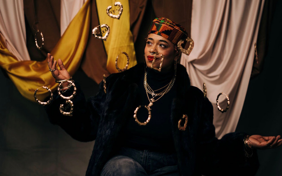 Darling Nikki wearing a Kente cloth pillbox hat, layered gold chains, and a black faux fur coat against draped gold fabric and suspended gold earrings by Everythang Dope.everythang dope vintage streetwear, vintage black fur coat outfit kente cloth hat vintage style, gold bamboo hoop earrings vintage, retro 90s streetwear lookbook, atlanta vintage clothing shop