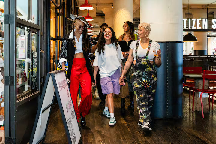 Four women walking hand-in-hand down a hallway in a restaurant or cafe, showcasing diverse fashion styles including bright red pants, a white t-shirt and purple shorts, and floral tie-dye overalls.