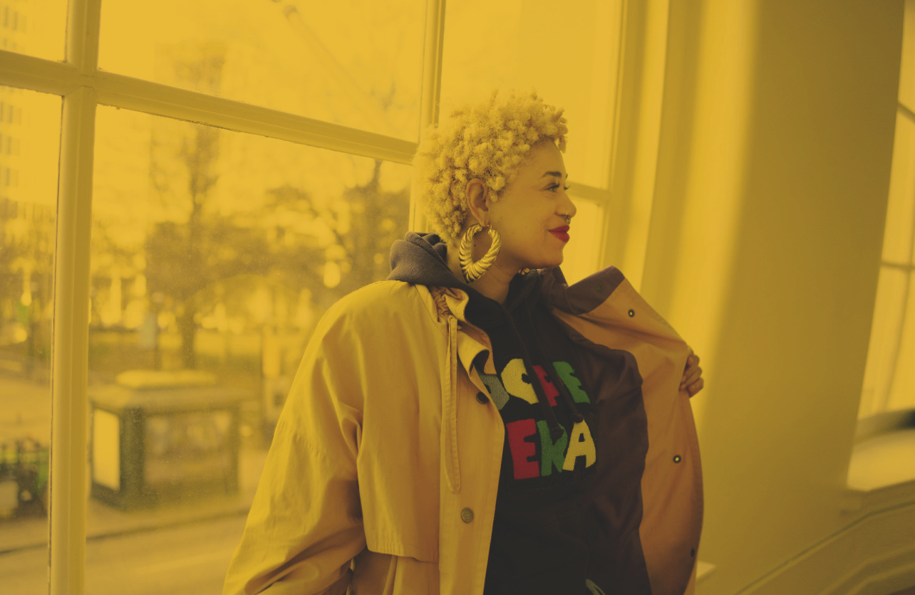 Vintage Darling Nikki with blond afro hair wearing gold bamboo hoop earrings, a gray hoodie, and a jacket backlit by a warm yellow tint inside a shop by Everythang Dope
everythang dope vintage streetwear, vintage streetwear lookbook
